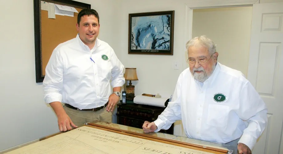 Jason Chiniche and his father, company founder James J. Chiniche, standing over a large survey drawing at the firm's drafting table, both wearing white shirts with the Chiniche logo.