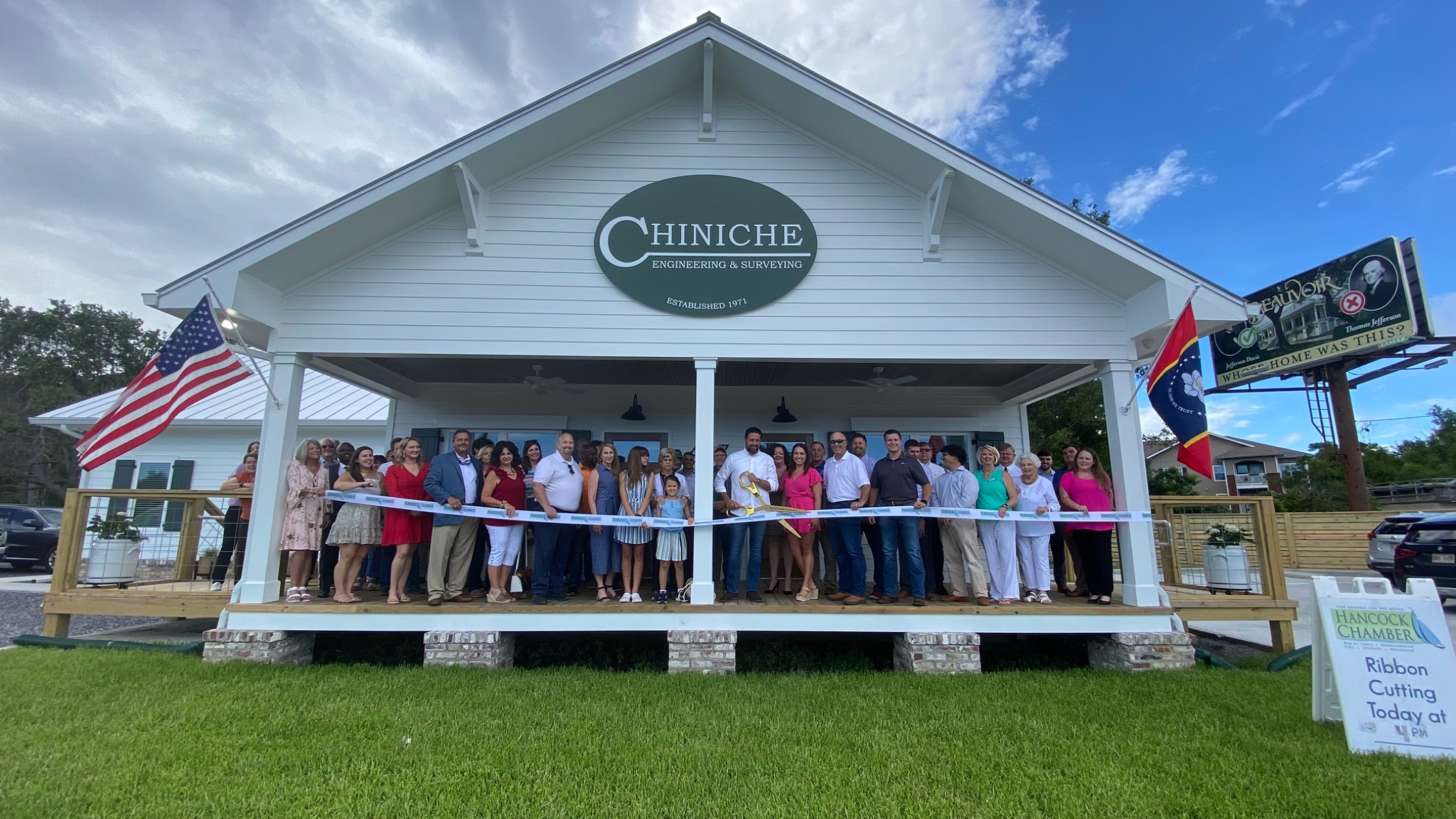 The Chiniche Engineering & Surveying team and guests hold a ribbon across the porch of the firm's new white Highway 90 office, beneath the redesigned green-and-white oval Chiniche logo sign.